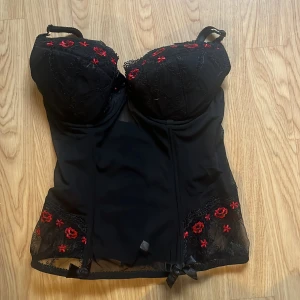 Svart korsett med röda broderade blommor - Säljer en svart korsett med vadderade kupor och justerbara axelband. Dekorerad med röda broderade blommor och spetsdetaljer upptill och nertill. Stängs med hyskor i ryggen. Perfekt för att skapa en snygg siluett.