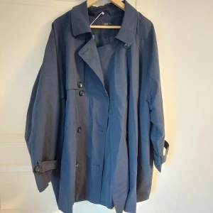 Blå trenchcoat från xlnt - Säljer en blå trenchcoat från xlnt i storlek 3XL. Jackan har klassisk krage, knappar framtill och bälte i midjan. Ärmarna har knäppning vid handlederna för extra detalj. Perfekt för dig som vill ha en stilren och tidlös jacka.