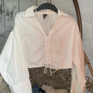Vit croppad skjorta med knyt - Säljer en vit croppad skjorta från H&M med lång ärm och klassisk krage. Skjortan har knytning framtill och en lätt v-ringning. Perfekt för en trendig och avslappnad stil.