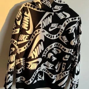 Svartvit hoodie från Nike med logga och text - Nike hoodie i svart och vitt med stora tryck av Nike-loggan och texten 'Just do it' över hela tröjan. Mjuk insida och huva. Perfekt för dig som gillar streetwear och vill sticka ut med en bold look.