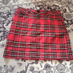 Röd rutig kortkjol med bälteshällor - Säljer en snygg röd och svart rutig kortkjol med bälteshällor och dragkedja fram. Kjolen är använd 1 gång, köpt från SHINE I storlek S 