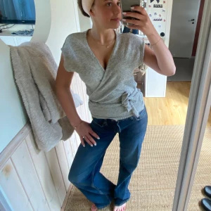 Grå omlottopp med knyt - Säljer en ljusgrå omlottopp med kort ärm och knyt i midjan. Toppen har v-ringning och är i mjukt material, perfekt för en avslappnad stil.