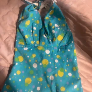 Turkos halternecktopp med prickigt mönster från Liss - Säljer en turkos halternecktopp från Liss med färgglatt prickigt mönster i gult, vitt och grönt. Toppen har knyt i nacken och  v-ringning framtill samt är i 80-tals stil