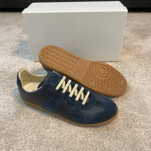 Maison Margiela Gats - Snygga mörkblå sneakers från Maison Margiela med beige sula och vita snören. Skorna har en mix av mocka och skinn, klassisk design och rund tå. Perfekta för dig som gillar stilrena och trendiga sneakers. Box, dustbags och kvitto ingår.
