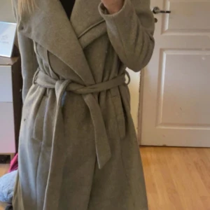 Grå kappa med bälte från ONLY - Säljer en stilren grå kappa från ONLY i storlek S. Kappan har ett brett slag, knytskärp i midjan och långa ärmar. Perfekt för dig som vill ha en klassisk och tidlös look.