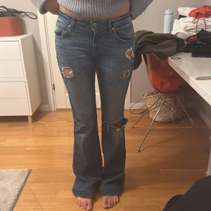 Blå bootcut jeans med slitningar - Snygga blå bootcut jeans med slitningar på låren och knäet. Klassisk femficksmodell med knapp och dragkedja framtill. Jeansen har hög midja och är i ett mjukt denimtyg som ger en avslappnad look.