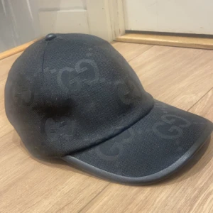 Svart keps från Gucci med GG-mönster - Säljer en svart keps från Gucci med diskret GG-mönster över hela kepsen. Kepsen har böjd skärm och justerbart spänne bak i skinn. Klassisk och stilren design med märkets logga invävd i tyget.