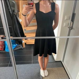 Svart klänning med breda axelband - Säljer en svart klänning med breda axelband och hjärtformad urringning. Klänningen har en markerad midja och utställd kjol i strukturerat tyg. 