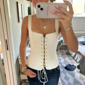 Vit korsett-topp med snörning - Säljer en vit korsett-topp med tunna vertikala ränder och snörning framtill. Toppen har breda axelband och en figurnära passform. Perfekt att styla med jeans eller kjol för en trendig look.
