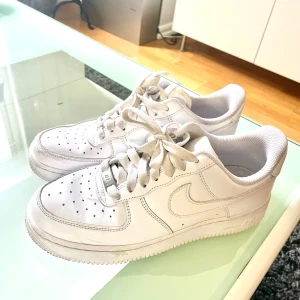Vita Nike Air Force 1 sneakers - Klassiska vita Nike Air Force 1 sneakers med rund tå och snörning. Skorna har en platt sula och är tillverkade i skinn med diskreta detaljer. Perfekta för en stilren och sportig look.
