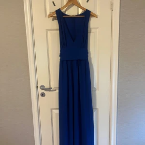 Blå långklänning med v-ringning - Säljer en elegant blå långklänning med djup v-ringning och bred midjedetalj. Klänningen är ärmlös och har en stilren, klassisk design som passar perfekt till bal eller fest. Dragkedja i ryggen för enkel påklädning.