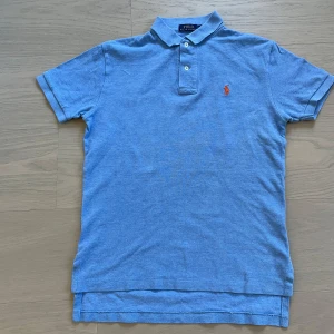 Blå pikétröja från Polo Ralph Lauren - Snygg blå pikétröja från Polo Ralph Lauren med klassisk krage och knappar framtill. Tröjan har korta ärmar och den ikoniska broderade loggan i orange på bröstet. Perfekt för en stilren och avslappnad look. Nypris 1000