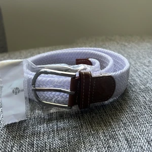 Vitt flätat bälte  - Snyggt vitt flätat bälte med detaljer i brunt skinn och silvrigt spänne. Perfekt accessoar för att lyfta din outfit. Bältet har klassisk design och passar till många olika stilar.