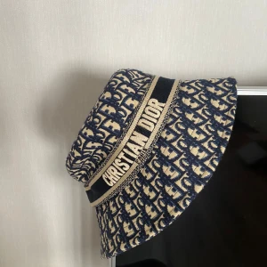  bucket hat från Christian Dior - Säljer en exklusiv bucket hat från Christian Dior med blått och beige mönster samt tydlig logga runt hatten. Hatten har ett klassiskt snitt och är tillverkad i ett tyg med struktur. Perfekt accessoar för att lyfta din stil. Oanvänd