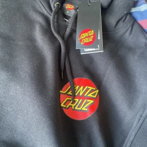 Svart Santa Cruz hoodie med tryck - Svart hoodie från Santa Cruz med klassiskt rött och gult logotryck på bröstet och ryggen. Tröjan har huva med dragsko och en stor magficka. Perfekt för dig som gillar streetwear och skate.