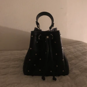 Svart väska med silvriga detaljer - Säljer en jätte snygg väska med silvriga prickar på. Den är i perfekt storlek. Medföljer band så man kan ha den över axeln istället för att hålla den. Den är i väldigt bra skick.