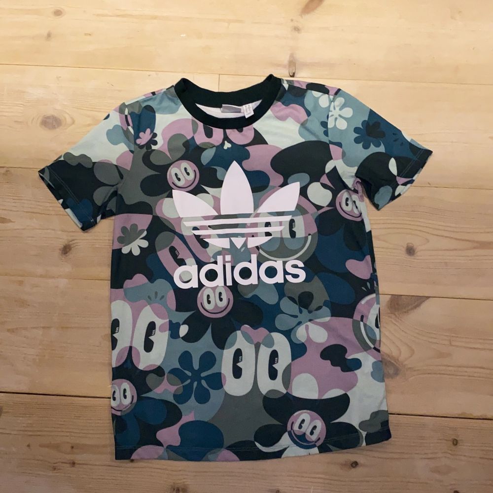 Sportig och cool mönstrad adidas t-shirt. 💚💙🖤💜🤍 Med stort adidastryck i vitt, mönstrad med blommor smileys och typ kamouflage, svart krage, och passar bra till träning, men kan även användas till vardags. 100% polyester. Mycket bra skick då den endast är använt fåtal gånger. XS men sitter lite oversized och snyggt på mig som är en XS/S. Funkar somsagt bra till träning och lek. DM vid intresse/fler frågor/ fler bilder.❣️(Det kan du göra här under för det står ”kontakta”) Avhämtning på Södermalm eller frakt till självkostnadspris.:). T-shirts.