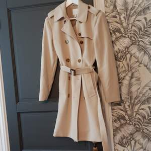 Beige trenchcoat från h&m med många snygga detaljer som dubbelknäppning med snygga knappar,   fickor knäppning vid kragen, skärp med snyggt spänne, slits bak mm. Oanvänd.