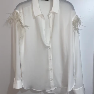 Tunn skjorta/blus från Zara - En vit, tunn skjorta i blusmaterial från Zara. Den har vita fjädrar på båda axlarna/överarmarna. Väldigt fin, passar även xs.