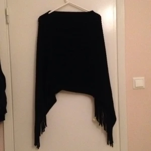  - Svart poncho med flätor längst ner. Kan skickas mot fraktkostnad.