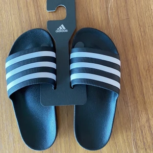 Adidas-slippers, tofflor, stl 38 - Oanvända Adidas tofflor i stl 38. Helt i plast. Nypris 199kr. Kan postas mot fraktkostnad på 55kr.