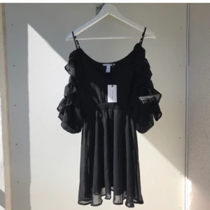 Klänning från Nelly - Så snygg svart klänning i chiffong med volanger och öppna armar. Aldrig använd med tags kvar. Passa på att fynda till billigt pris! Passar även S.