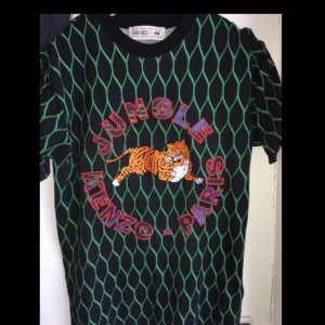  - En svart Kenzo tröja med grönt ”galler”, en broderad tiger och texten ”JUNGLE KENZO-PARIS” broderat på framsidan. Tröjan är från KenzoXh&m kollektionen. Använt max 3 gånger och säljer för att jag inte använder den längre. Köparen står för frakt. 