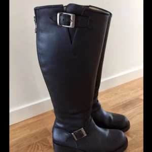  - Skinn boots med dekorrem med spänne i antik gulmetall över vristen från Johnny Bulls. Slits upptill på skaftet med reglerbar rem över, så skaftvidden kan regleras något. Dold resårkil på skaftets insida. Metalldragkedja mitt bak på skaftet.   Skafthöjd: ca 42 cm Färg: Svart   Använda en gång så skorna är i mycket fin skick. Nypris 1899:-