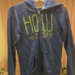 Hollister kofta - Blå kofta från hollister med coolt tryck. Tröjan är jättemjuk pch skön med fickor. Den har ett blixtlås!