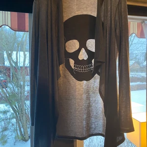 Grå kofta med svartglittrig dödskalle på ryggen - Snygg kofta med volang framtill och en svartglittrig dödskalle på ryggen. Endast använd ett fåtal gånger, är i nyskick.