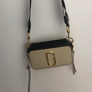 Marc Jacobs snapshot-bag - En snygg mindre crossbody väska i hårt läder vit och svart med guldiga detaljer. Väskan är uppdelad i två fack. Jag har använt den en del så axelbandet är lite slitet och de guldiga detaljerna är lite rostiga. Väskan köptes för 3000kr men pågrund av att den är sliten säljer jag den för 1/3 av nypriset🤓