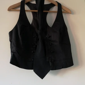 Svart figursydd väst med slips - Svart figursydd väst. Svart slips ingår. Snygg med tex vit t-shirt. Knäppning fram med hakar. Påsydda knappar. 