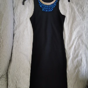 Svart knälång tajt klänning strl 38 - Klänning använd en gång, figurformad och strecthig i sin material. Fin detalj vid nackkragen med halsband fastsydd (går nog att ta bort om så önskas) strl mellan S/M (38)