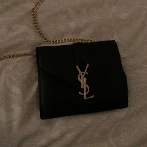 Ysl väska - Säljer denna superfina väskan (vet ej om den är äkta) från Ysl! I en fin läderimitation med guldiga detaljer. Det går att vika in kedjan om man vill ha den kortare, superfint blir den som axelväska! Buda från 100kr