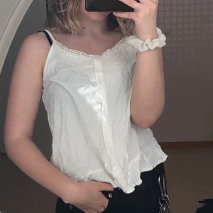 Vitt linne med volanger  - Ett vitt gulligt linne med volanger längst ner och knappar längst fram på tröjan som går neråt. Är i bra skick eftersom jag aldrig har burit det. Frakten ingår inte i priset