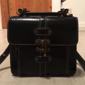  - Liten snygg retroväska i skinn med antika detaljer och axelband. Finns även ett litet handtag och man väljer att ta bort axelbandet. Perfekt att ha sina viktigaste tillhörigheter i nu när sommaren närmar sig och klänningen åker på! Finns att hämta i stockho