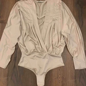  - V-ringad body i silkes material från NELLY i storlek XS. Nästan helt oanvänd! På bilden ser den ut att vara mer beige, men skulle säga att den är mer i en rosa nyans i verkligheten. Frakt tillkommer ♥️