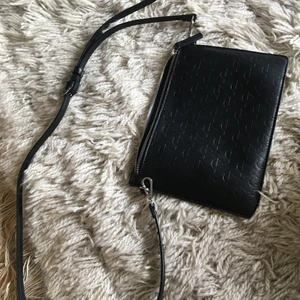 Carin Wester axelremsväska  - CW axelremsväska i svart läderliknande material, smalt reglerbart axelband med spänne. Mått 19 x 26 cm. Ett innerfack med dragkedja plus två extra fack. 