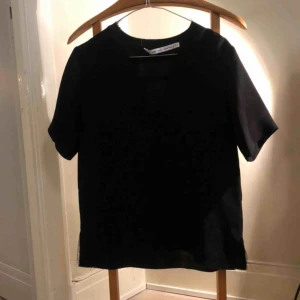  - Svart blus/topp,100% bomull, med v-ringning och slitsar i sidan. Kanterna är avklippta (så är designen) därav hänger några trådar löst men annars i bra skick.  Kan skickas mot fraktkostnad eller mötas i sthlm. 