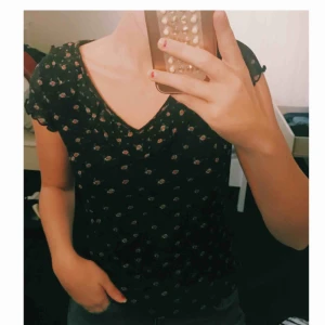  - T-shirt med små blommor på. Det är volanger uppe vid halsen. 