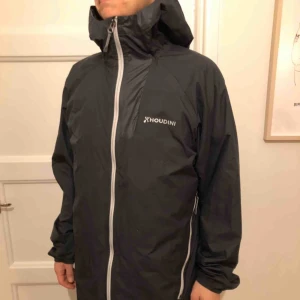  - Fin Houdini vindjacka, perfekt lite längre parkasmodell, vind- och vattenavstötande. Lätt och packar, kan packas ned i sin egen innerficka. Fint begagnat skick