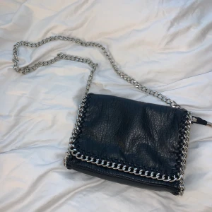 Axelremsväska - Snygg, svart axelremsväska med silvriga detaljer. Från märket TIAMO och köpt på Scorett. I mycket bra skick. Pris 150kr. Frakt tillkommer.