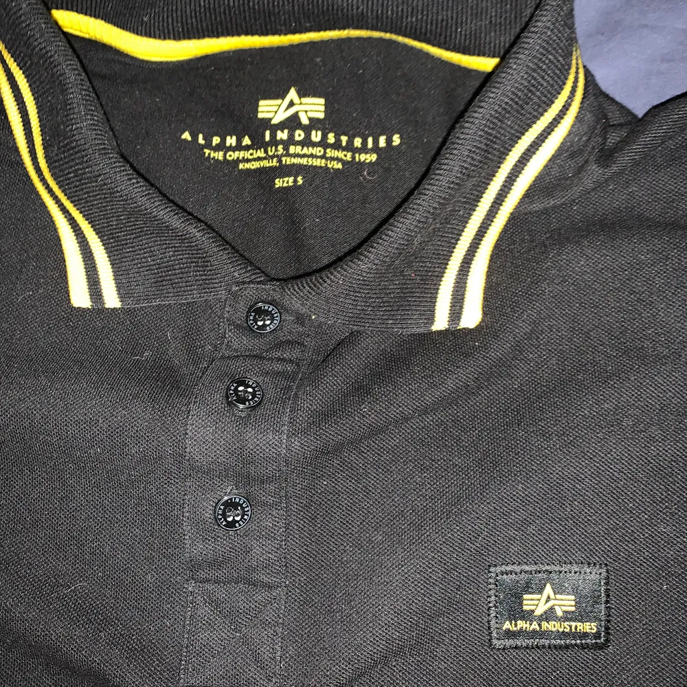 Svart Piké T-shirt med gula detaljer från Alpha Industries köpt 500kr från Zalando säljes för 150kr.. T-shirts.