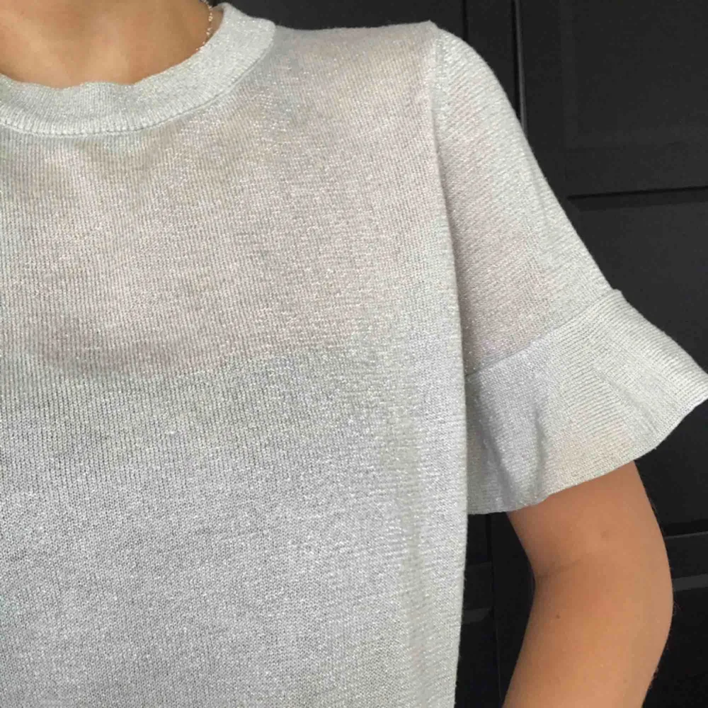 En ljusgrå glittrig t-shirt med volangdetalj vid ärmarna och nedre kanten✨ så snygg passform. Toppar.