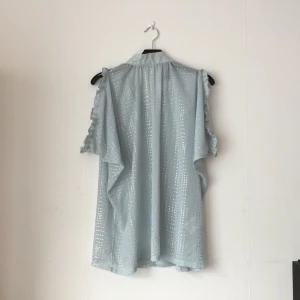  - Ljusblå sommarblus/top för sena bar/klubbkvällar med silvermönster och vida öppna ärmar med volang. Knyts med band i nacken, generös vidd så funkar nog för en medium också. Nyskick. Mer ljusblå än bilderna visar (molnig dag). 