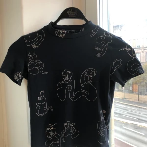 Mönstrad T-shirt med polo  - SÅ fin t-shirt från Monki med ett coolt mönster av ansiktsmotiv. Tröjan är marinblå. Halvpolo. Materialet är stretchigt vilket gör att tröjan sitter galet snyggt på. Har massor av snygga toppar uppe på min profil - kombinera gärna fraktpriset genom att köpa andra plagg. 🤩