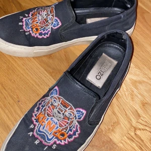 Kenzo slip ons - Säljer mina svarta Kenzo slip ons, dem är använda väl men har fortfarande mycket att ge. Tagits hand om väl! Inget slitage på skon någonstans endast ”veck”. Storlek 38. Det är äkta och är köpta på Kenzo original sida. Nypris ca 1450kr. (Ska tvättas innan jag skickar dem såklart!)