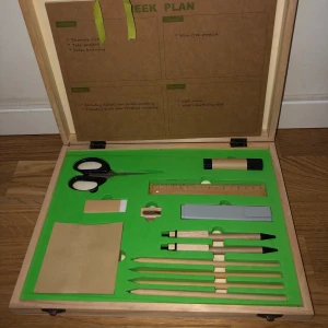 Väska med kontorstillbehör - En träväska som innehåller 2 stiftpennor, 4 blyertspennor, gul highlightpenna, 15 cm lång linjal, limstift, sax, sudd, pennvässare och post-it lappar. 