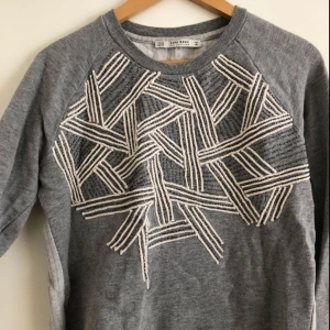  - Grå långärmad tröja i lite tjockare bomullstyg. Broderat mönster med pärlor på framsidan. Större än vanlig XS i storleken.
