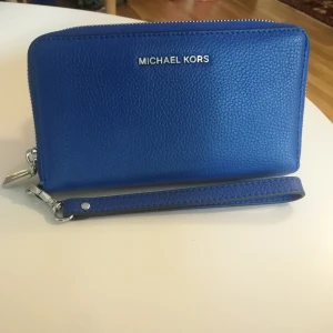  - Michael Kors plånbok, ny, oanvänd med samtliga lappar. Finns i söderort.  Modell: Mercer Flat Case  Mått: Bredd ca 20 cm Höjd ca 10,5 cm Djup ca 2 cm Färg: Blå/electric blue med silver detaljer. Storlek: One size 100% Skinn Foder: Textil Gratis leverans i söderort. 